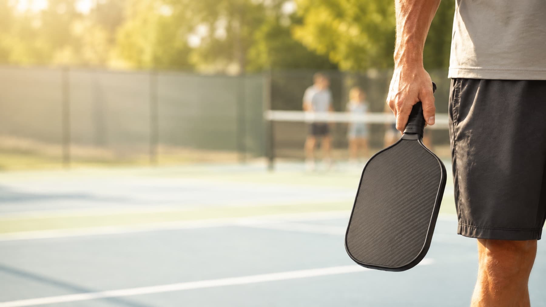 A pickleball paddle held at a player's side on a sunlit outdoor court.